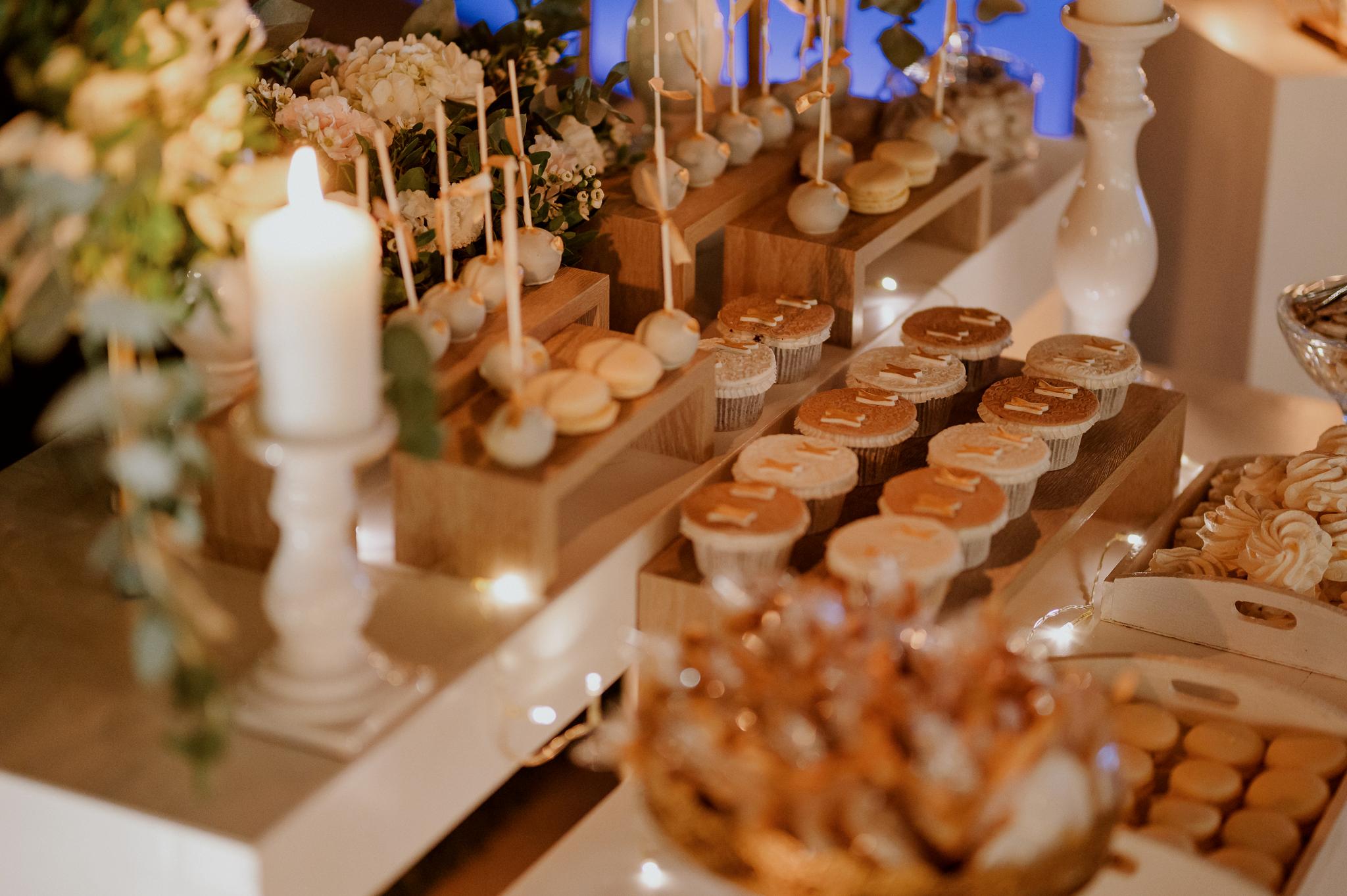 candy bar κοντινή λήψη σε γλυκά τα οποία είναι cup cake και macaron με τα αρχικά του ζευγαριού από πάνω σε ζαχαρόπαστα σε χρώματα λευκά ροζ και χρυσά . Επάνω στο τραπέζι έχει cake pops όπως επίσης και στολισμό ο οποίος είναι λουλούδια λευκά τριαντάφυλλα