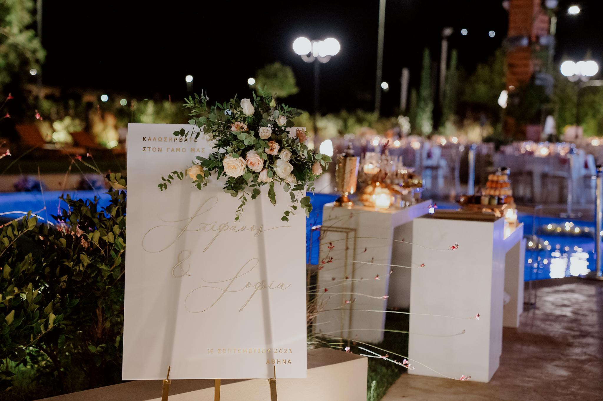 Κοντινή φωτογραφία από την σήμανση που βρίσκετε στην είσοδο του κτήματος η οποία αναγράφει καλώς ήρθατε στο γάμο μας και το όνομα του ζευγαριού . Επάνω στην σήμανση αυτή βρίσκετε μια σύνθεση με λουλούδια τριαντάφυλλα λευκά και σομόν με πρασινάδες.