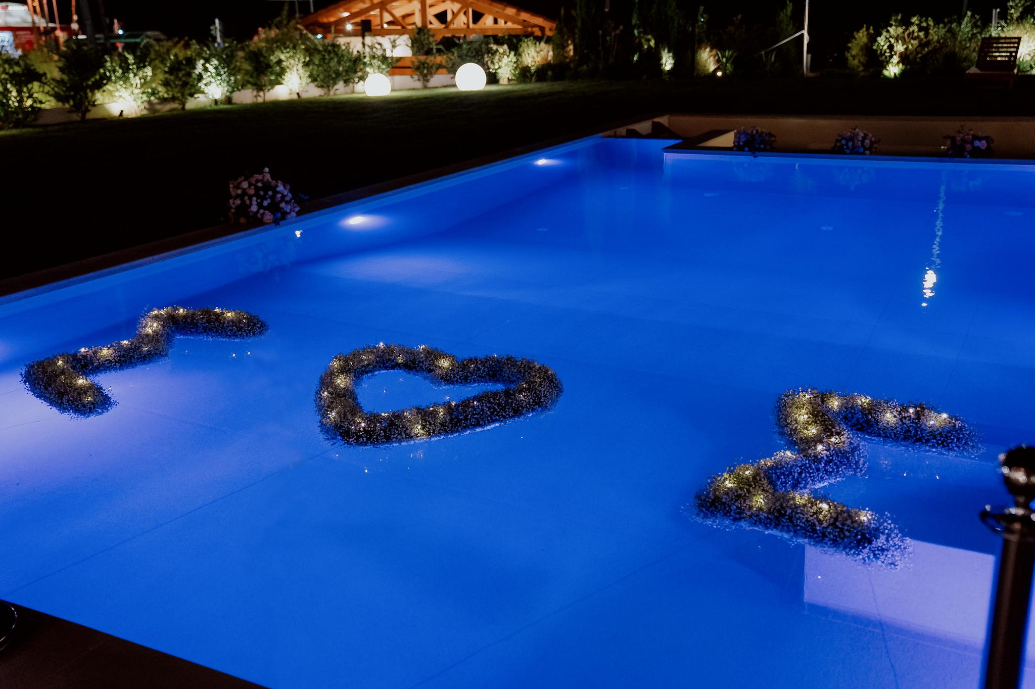Φωτογραφία που απεικονίζει την πισίνα του κτήματος και μέσα στην πισίνα έχουν βάλει τα αρχικά του ζευγαριού από λουλούδια με μια καρδιά στην μέση. Είναι το γράμμα Σ με μια καρδία δίπλα και ξανά το γράμμα Σ