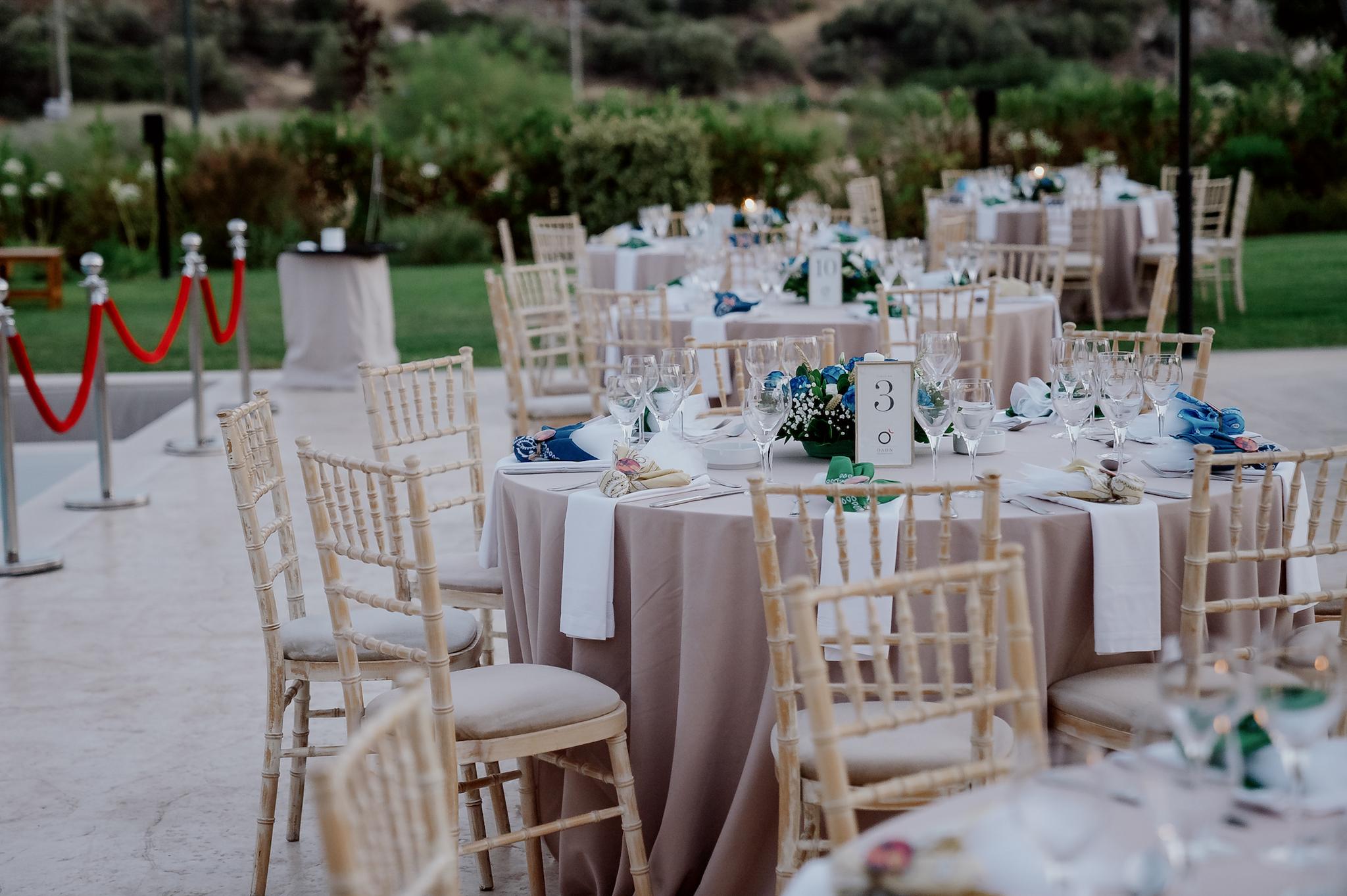Μακρινή φωτογραφία στα τραπέζια του κτήματος , λουλούδια βρίσκονται επάνω στα τραπέζια τα οποία περιέχουν μπλε τριαντάφυλλα και λευκά . Επάνω στο κάθε τραπέζι βρίσκονται τοποθετημένες και η μπομπονιέρες