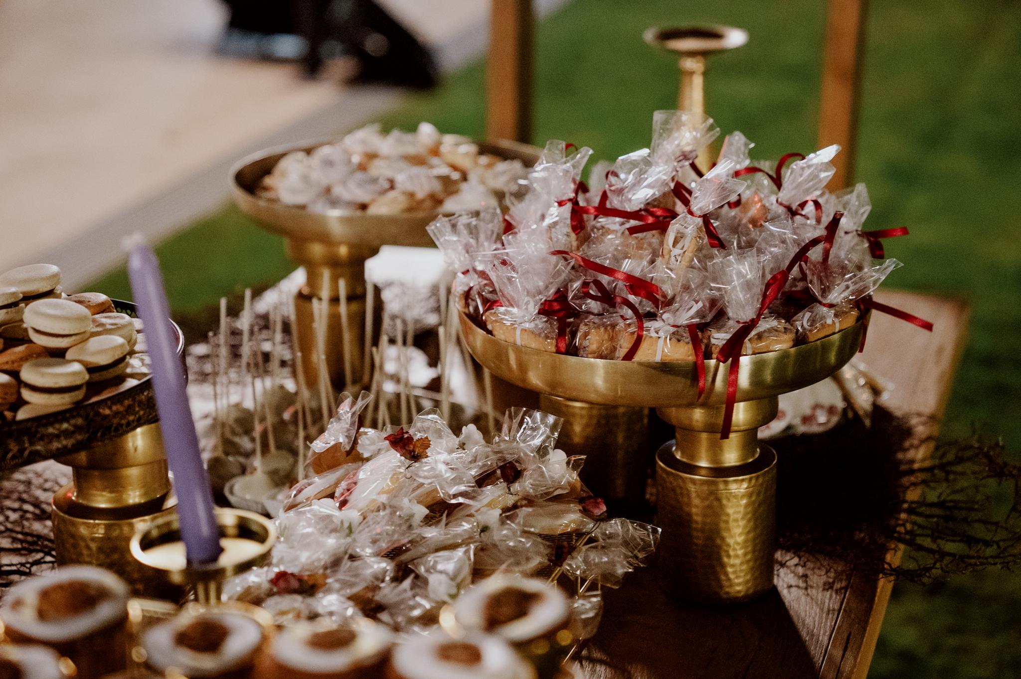 Με φόντο τον πράσινο κήπο και της επιδαπέδιες φωτιζόμενες μπάλες φαίνεται το candy bar το οποίο είναι σε χρωματισμούς του χρυσού και επάνω στα χρυσά σκεύη βρίσκονται cookies μαζί με μαρέγκες και cake pops