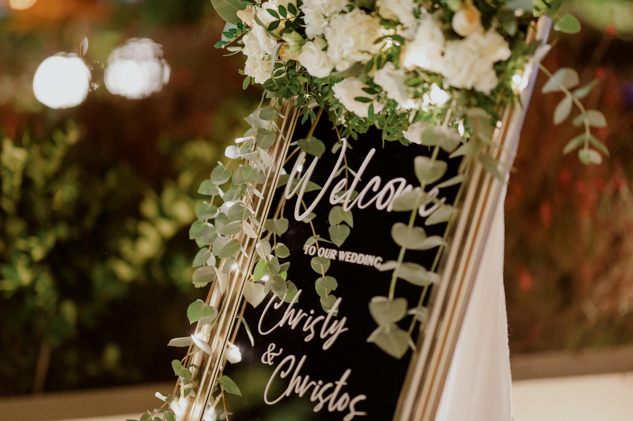 Κοντινή φωτογραφία από την σήμανση που βρίσκετε στην είσοδο του κτήματος η οποία αναγράφει καλώς ήρθατε στο γάμο μας και το όνομα του ζευγαριού . Επάνω στην σήμανση αυτή βρίσκετε μια σύνθεση με λουλούδια τριαντάφυλλα λευκά με πρασινάδες.