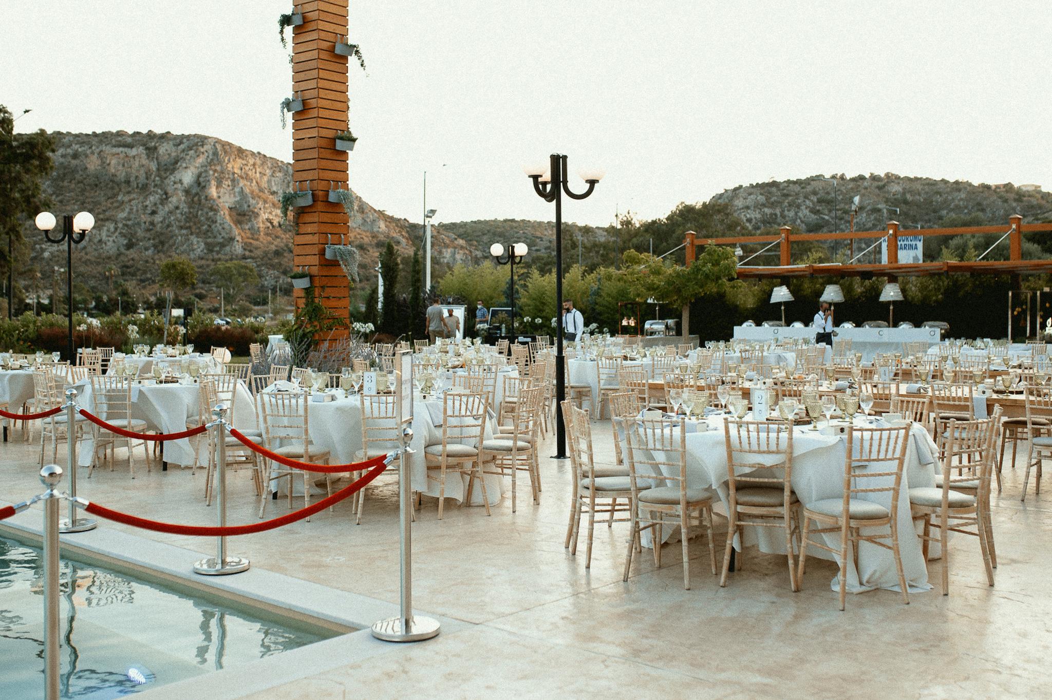 Απομακρυσμένη φωτογραφία με στρωμένες ροτόντες και μοναστηριακά τραπέζια. Περιμετρικά από τα τραπέζια είναι στημένες οι καρέκλες ξύλινες tiffany