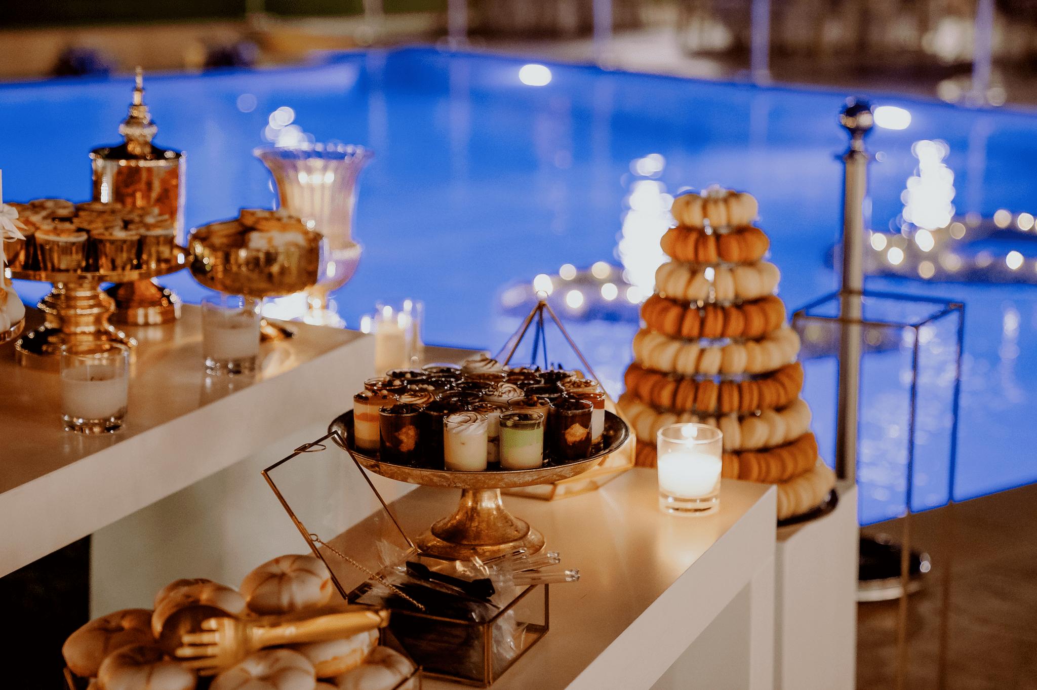 Φωτογραφία από το candy bar που απεικονίζει τα macarons σε ένα πύργο κεράκια όπως επίσης και ατομικά γλυκά μαρέγκες και μπισκότα με τα αρχικά του ζευγαριού. Στο βάθος φαίνεται η πισίνα.