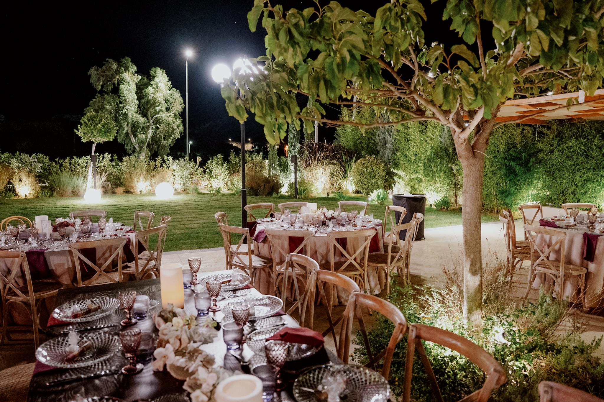 Βραδινή φωτογραφία με φόντο το πράσινο και και τα πολύχρωμα φυτά. Φαίνεται το μοναστηριακό τραπέζι αλλά και οι ροτόντες τραπέζια μαζί με τις ξύλινες χιαστί καρέκλες. Επάνω στα τραπέζια έχει σουπλά με τοποθετημένες μπομπονιέρες επάνω και πολύχρωμα ποτήρια του νερού και του κρασιού. Έχει στολισμό από λουλούδια και αχιβάδες σε μπορντό χρώμα. Στο φόντο υπάρχει το πράσινο γκαζόν και τα φυτά του κήπου.