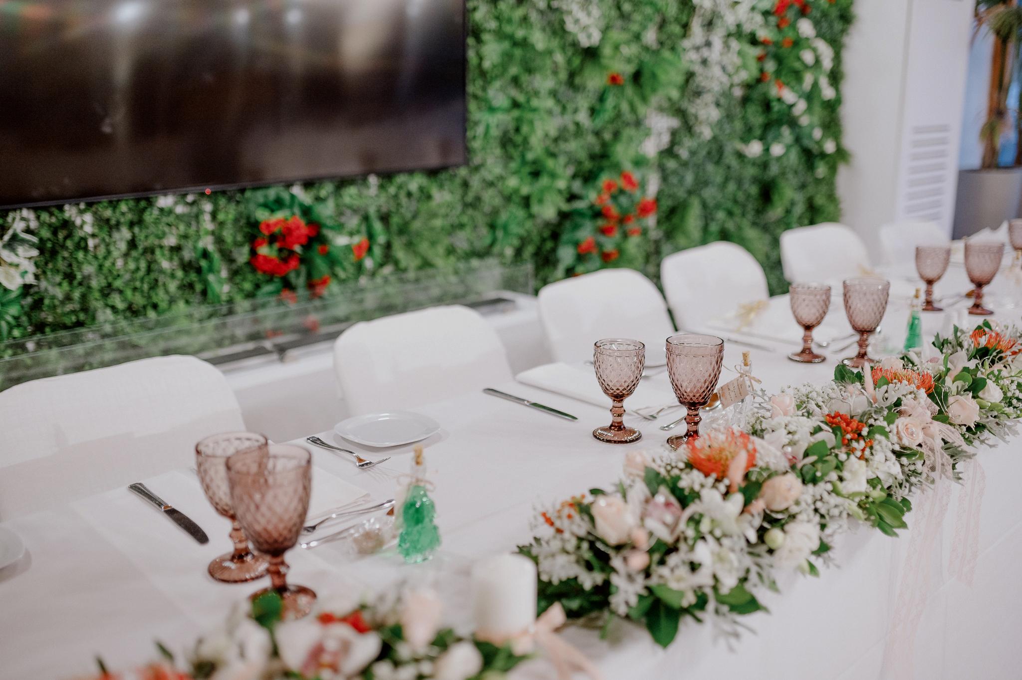 Κοντινή φωτογραφία από το νυφικό τραπέζι που φαίνονται τα λουλούδια τα οποία είναι τριαντάφυλλα σομόν ορτανσίες και πρασινάδα ενδιάμεσα. Δείχνει και τα ποτήρια τα οποία είναι σε ροζ χρώμα