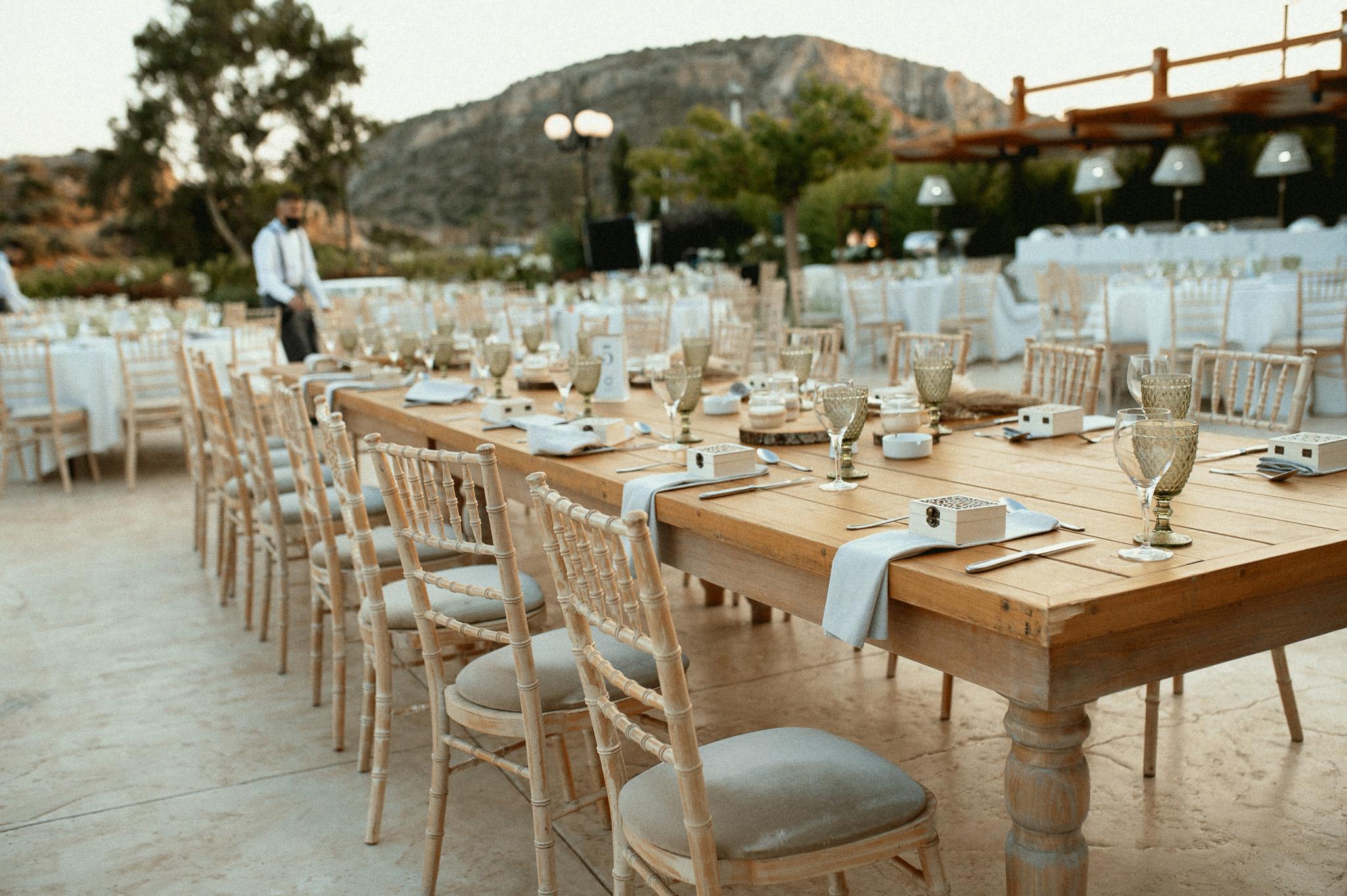 Κοντινή φωτογραφία σε μοναστηριακό τραπέζι με τοποθετημένες καρέκλες ξύλινες tiffany . Τα μοναστηριακά τραπέζια είναι στρωμένα και επάνω έχουν και τον στολισμό ο οποίο είναι ένα βάζο με Pampas σε διάφορα χρώματα