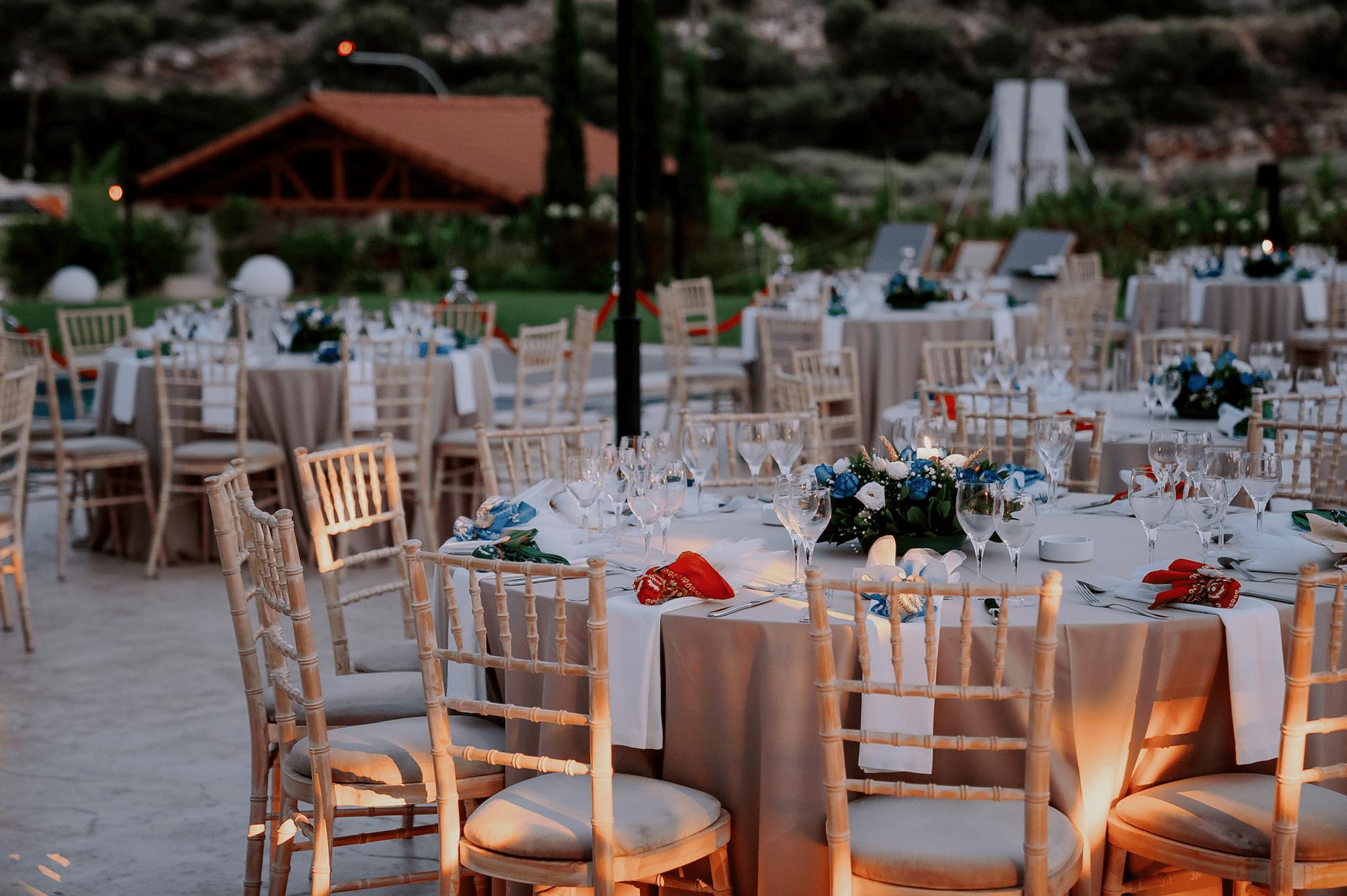 Μακρινή φωτογραφία στα τραπέζια του κτήματος , λουλούδια βρίσκονται επάνω στα τραπέζια τα οποία περιέχουν μπλε τριαντάφυλλα και λευκά . Επάνω στο κάθε τραπέζι βρίσκονται τοποθετημένες και η μπομπονιέρες