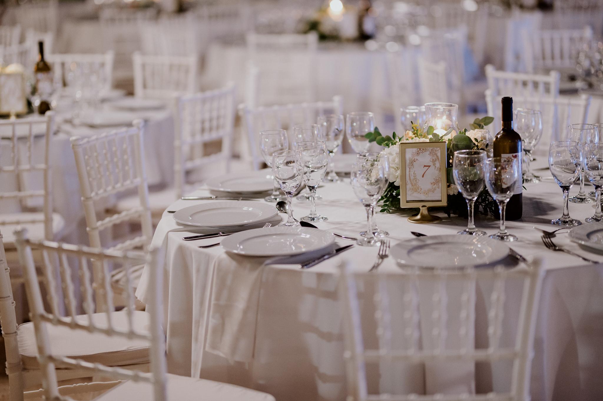 Κοντινή φωτογραφία με στρωμένα τραπέζια και καρέκλες περιμετρικά. Τα τραπέζια είναι στολισμένα με τριαντάφυλλα και ορχιδέες και λευκά τριαντάφυλλα με ένα αναμμένο κερί στην μέση . Στο φόντο έχει ένα πράσινο κήπο με γκαζόν.