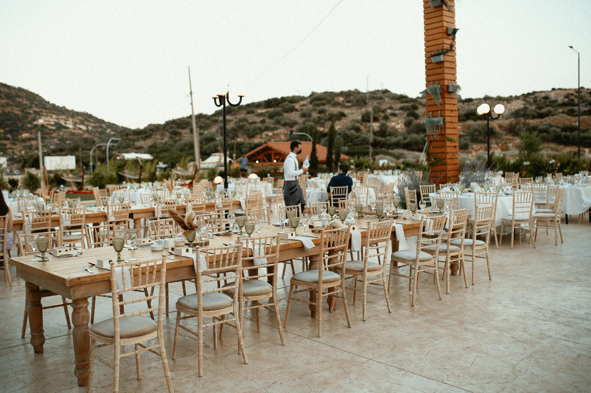 Απομακρυσμένα τραπέζια μοναστηριακά με tiffany καρέκλες περιμετρικά επάνω τα τραπέζια είναι στρωμένα από μαχαιροπίρουνα και ποτήρια σε χρώμα πράσινο.