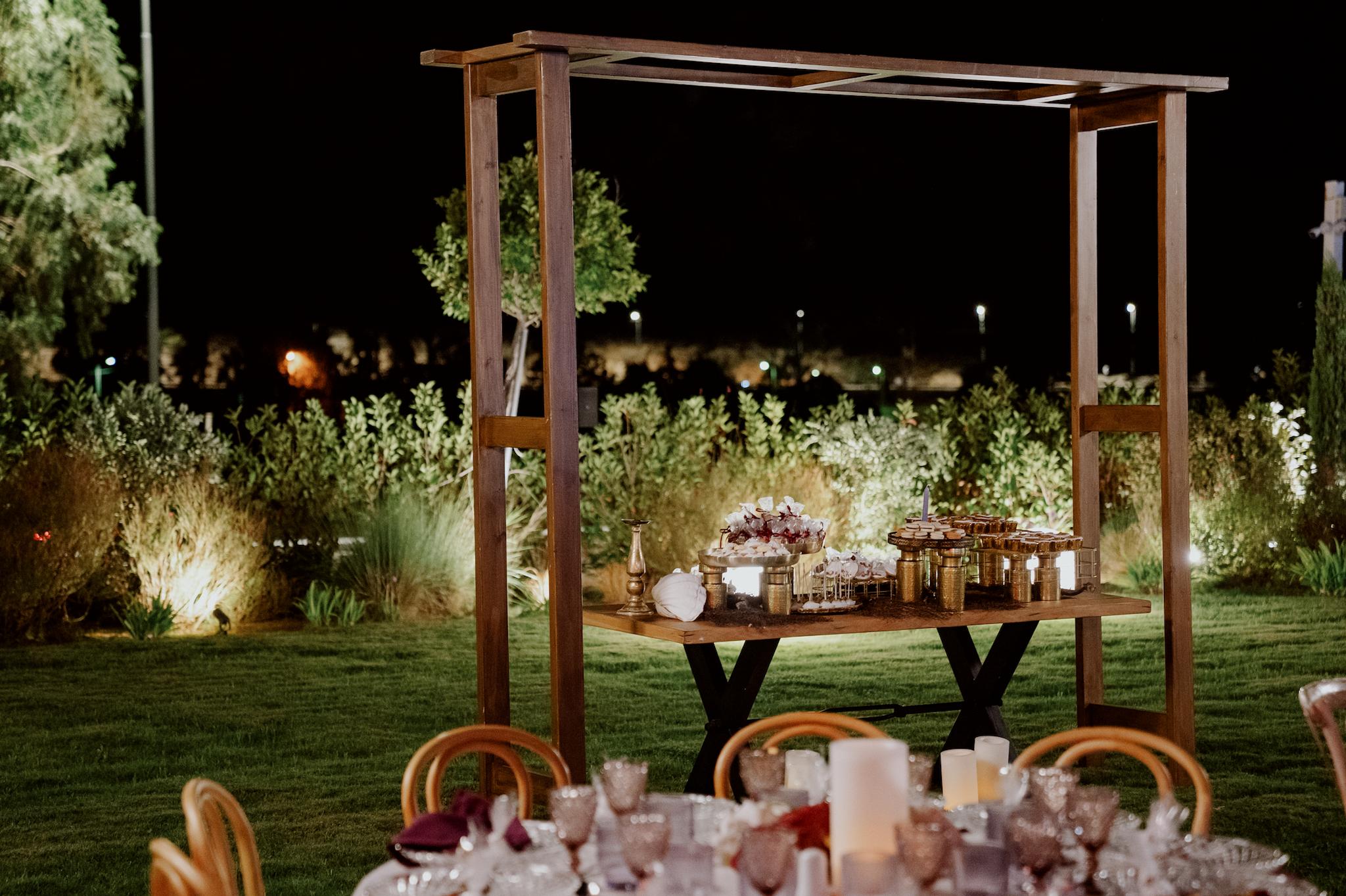 Με φόντο το πράσινο γκαζόν και τον κήπο με τα λουλούδια φαίνεται το candy bar με το μοναστηριακό τραπέζι. Επάνω στο τραπέζι βρίσκονται λεπτομέρειες χρυσές και επάνω σε αυτά τα γλυκά όπως cookies cake pops cup cake Και μαρέγκες