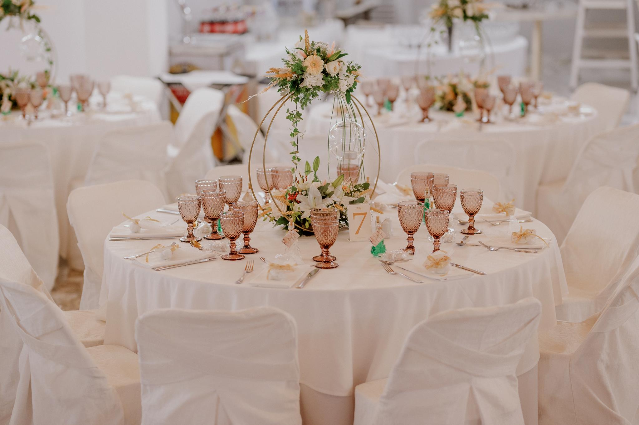 Φωτογραφία ροτόντας στον εσωτερικό χώρο του κτήματος με στολισμό του τραπεζιού με κερί να κρέμεται και στολισμό από ορχιδέα και τριαντάφυλλα σομόν με πράσινα φυτά. Στο τραπέζι βρίσκονται ποτήρια χρώματος ροζ και μαχαιροπίρουνα μαζί με συνοδευτικά γλυκά