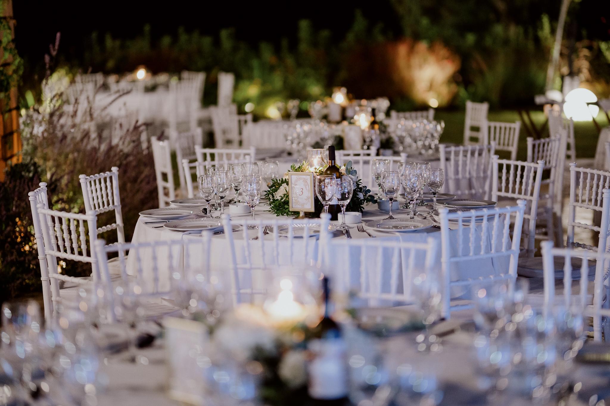 Απομακρυσμένη φωτογραφία με στρωμένα τραπέζια και καρέκλες περιμετρικά. Τα τραπέζια είναι στολισμένα με τριαντάφυλλα και ορχιδέες και λευκά τριαντάφυλλα με ένα αναμμένο κερί στην μέση . Στο φόντο έχει ένα πράσινο κήπο με γκαζόν.