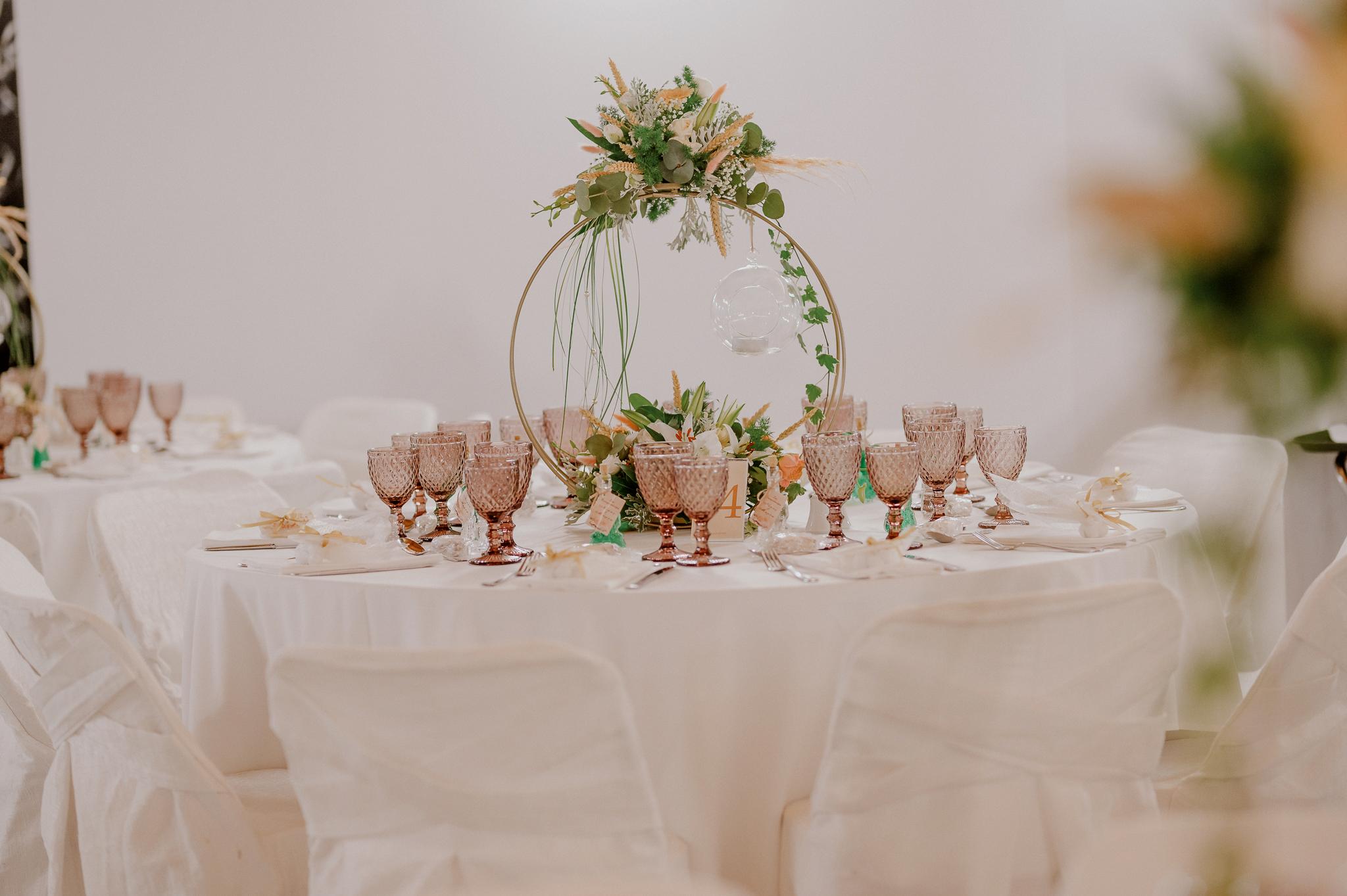 Φωτογραφία ροτόντας στον εσωτερικό χώρο του κτήματος με στολισμό του τραπεζιού με κερί να κρέμεται και στολισμό από ορχιδέα και τριαντάφυλλα σομόν με πράσινα φυτά. Στο τραπέζι βρίσκονται ποτήρια χρώματος ροζ και μαχαιροπίρουνα μαζί με συνοδευτικά γλυκά