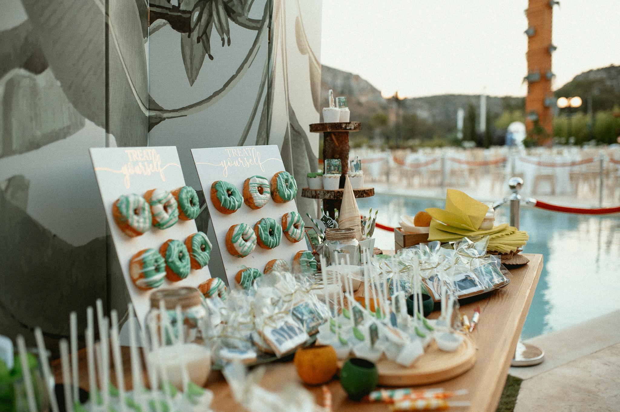 Πρωινή φωτογραφία από το candy bar επάνω σε μοναστηριακό τραπέζι. Με φόντο ένα πανί το οποίο έχει πράσινα φύλλα. Επάνω στο τραπέζι έχει donuts μαζί με cake pops cookies και cup cake