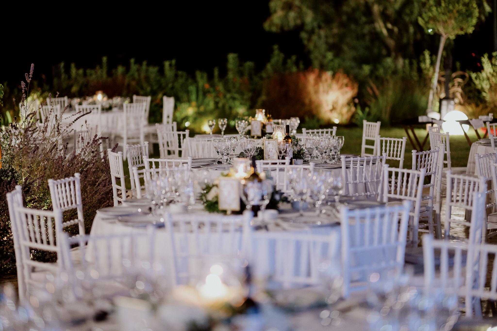 Απομακρυσμένη φωτογραφία με στρωμένα τραπέζια και καρέκλες περιμετρικά. Τα τραπέζια είναι στολισμένα με τριαντάφυλλα και ορχιδέες και λευκά τριαντάφυλλα με ένα αναμμένο κερί στην μέση . Στο φόντο έχει ένα πράσινο κήπο με γκαζόν.