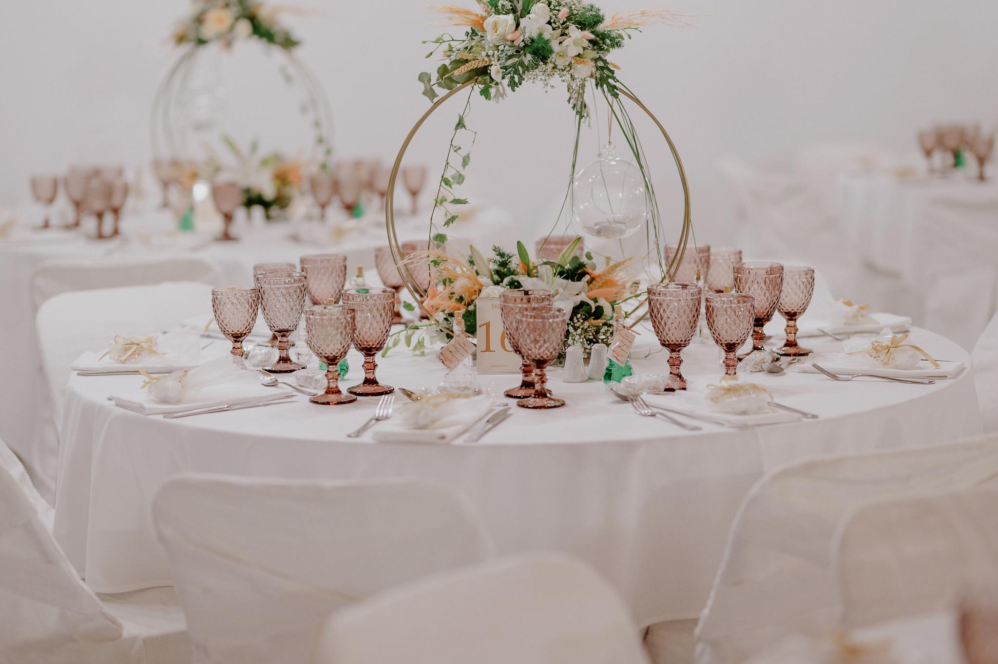 Φωτογραφία ροτόντας στον εσωτερικό χώρο του κτήματος με στολισμό του τραπεζιού με κερί να κρέμεται και στολισμό από ορχιδέα και τριαντάφυλλα σομόν με πράσινα φυτά. Στο τραπέζι βρίσκονται ποτήρια χρώματος ροζ και μαχαιροπίρουνα μαζί με συνοδευτικά γλυκά