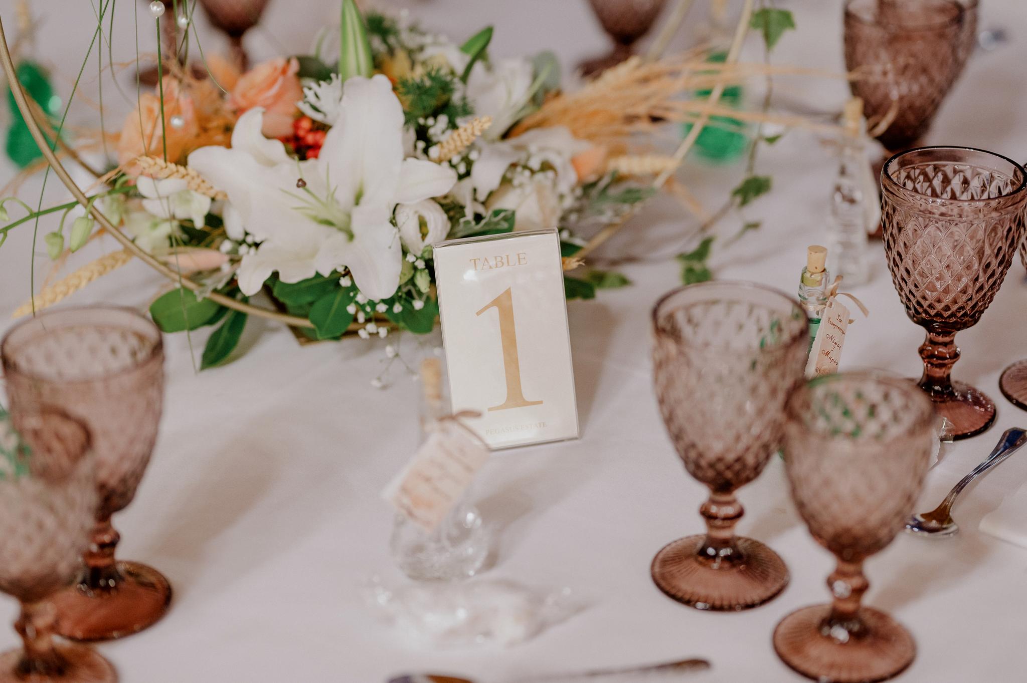 Κοντινή φωτογραφία με νούμερο τραπεζιού που φαίνεται και ο στολισμός από πίσω με ορχιδέες και Pampas και τριαντάφυλλα σομον. φαίνονται τα ποτήρια τα οποία είναι σε χρώμα ροζ