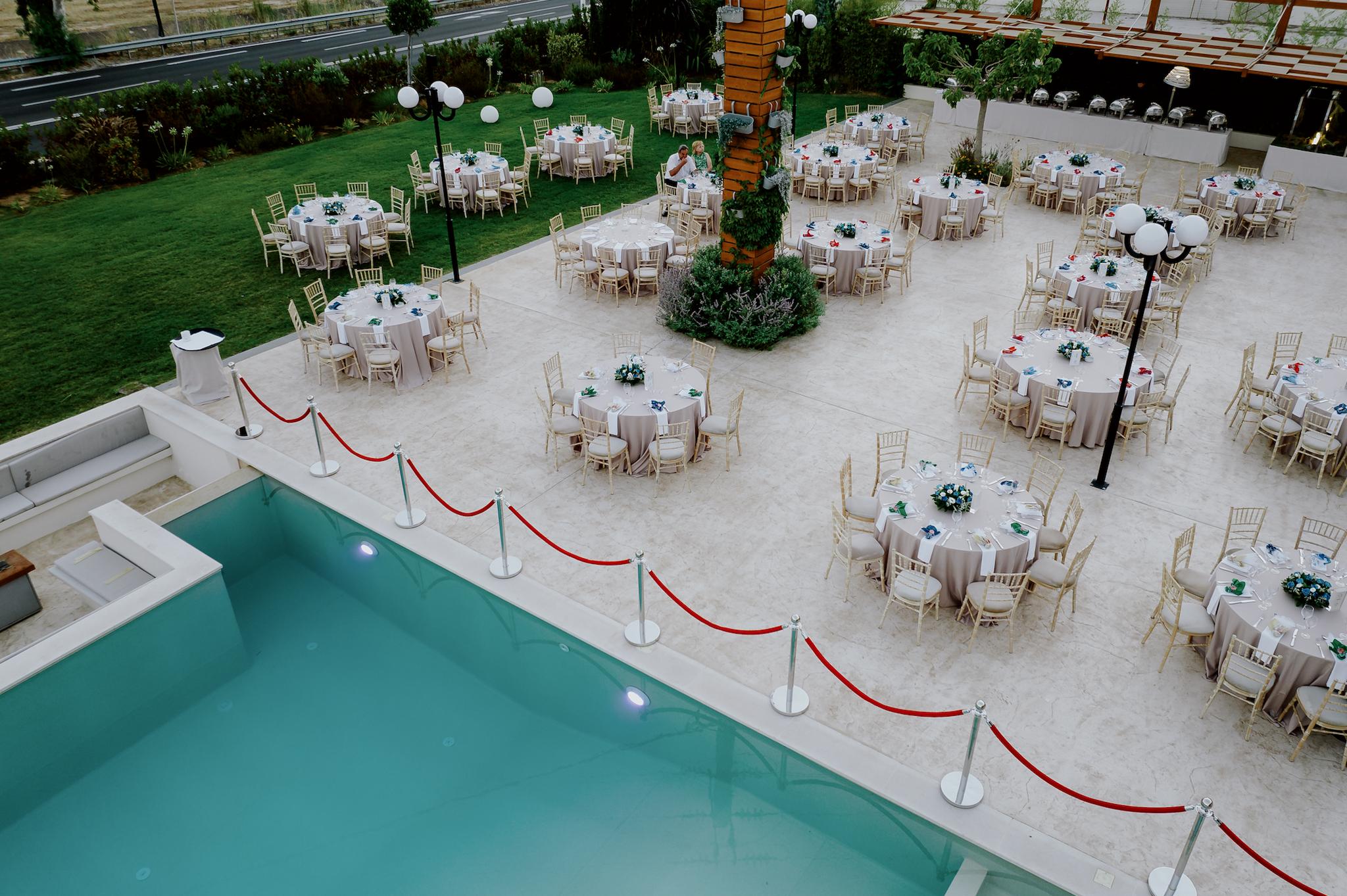 Πανοραμικό πλάνο σε πρωινή λήψη. Φαίνονται οι ροτόντες στρωμένες περιμετρικά με tiffany καρέκλες. Επάνω τα τραπέζια είναι στρωμένα και στο βάθος φαίνεται οι πισίνα του κτήματος όπως επίσης και ο πράσινος κήπος μαζί με το γκαζόν.