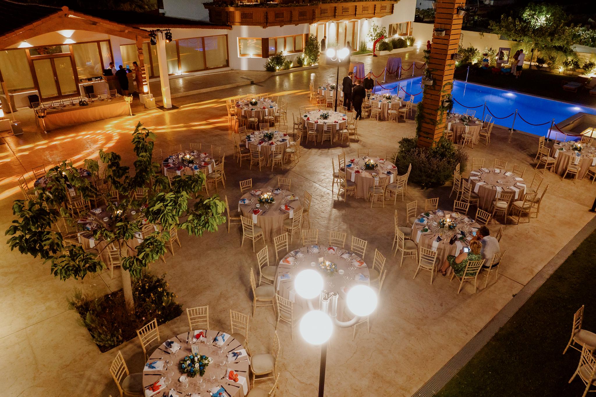 Πανοραμικό πλάνο σε βραδινή λήψη. Φαίνονται οι ροτόντες στρωμένες περιμετρικά με tiffany καρέκλες. Επάνω τα τραπέζια είναι στρωμένα και στο βάθος φαίνεται οι πισίνα του κτήματος