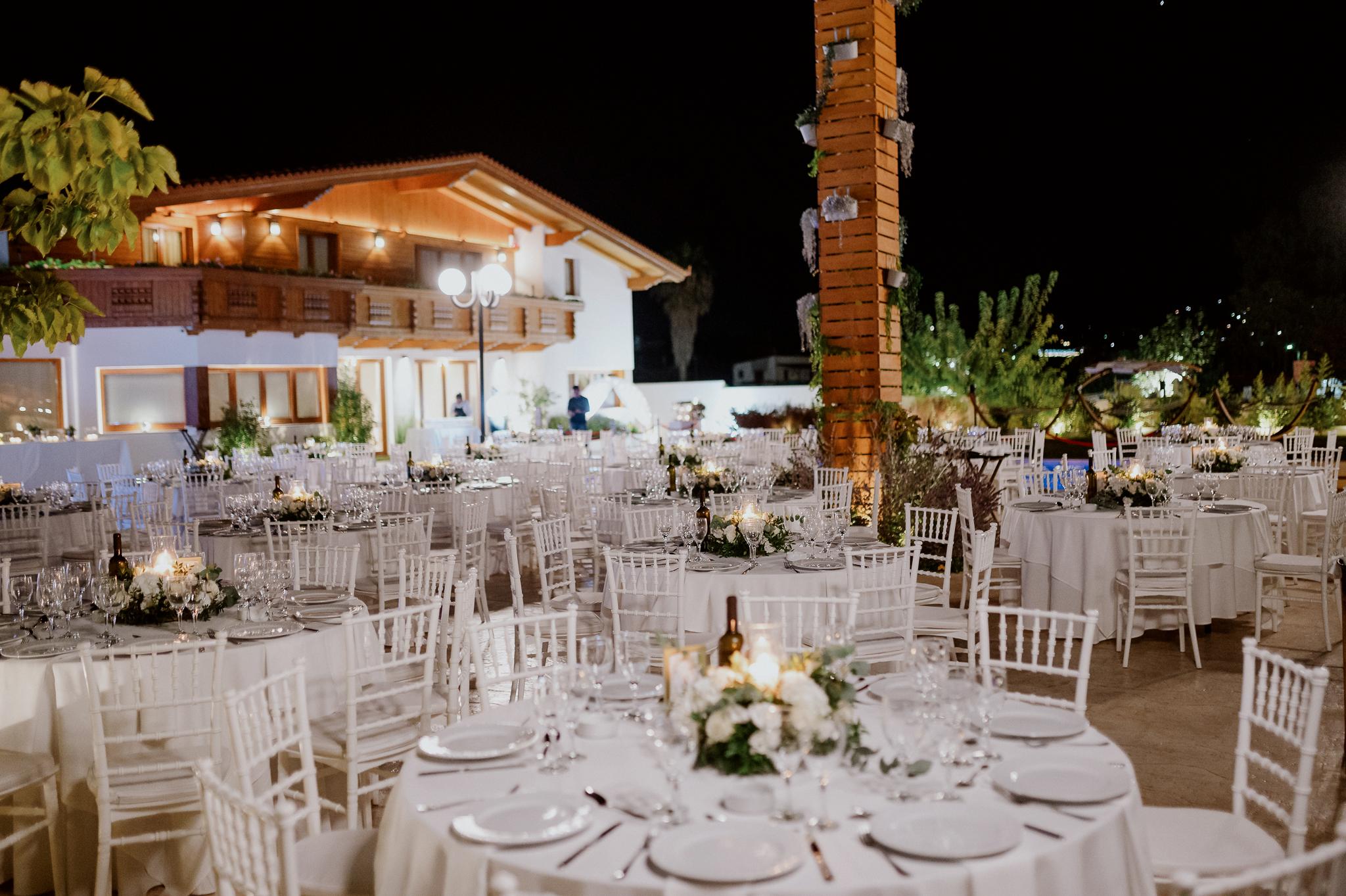 Με φόντο το ελβετικό σαλέ και τις ξύλινες λεπτομέρειες έχουν στρωθεί στον προαύλιο χώρο του κτήματος ροτόντες τραπέζια με περιμετρικά καρέκλες. Στην μέση του κάθε τραπεζιού βρίσκετε μια σύνθεση από ορχιδέες και λευκά τριαντάφυλλα. Επίσης βρίσκετε στην μέση του κάθε τραπεζιού βρίσκετε ένα μπουκάλι κρασί.