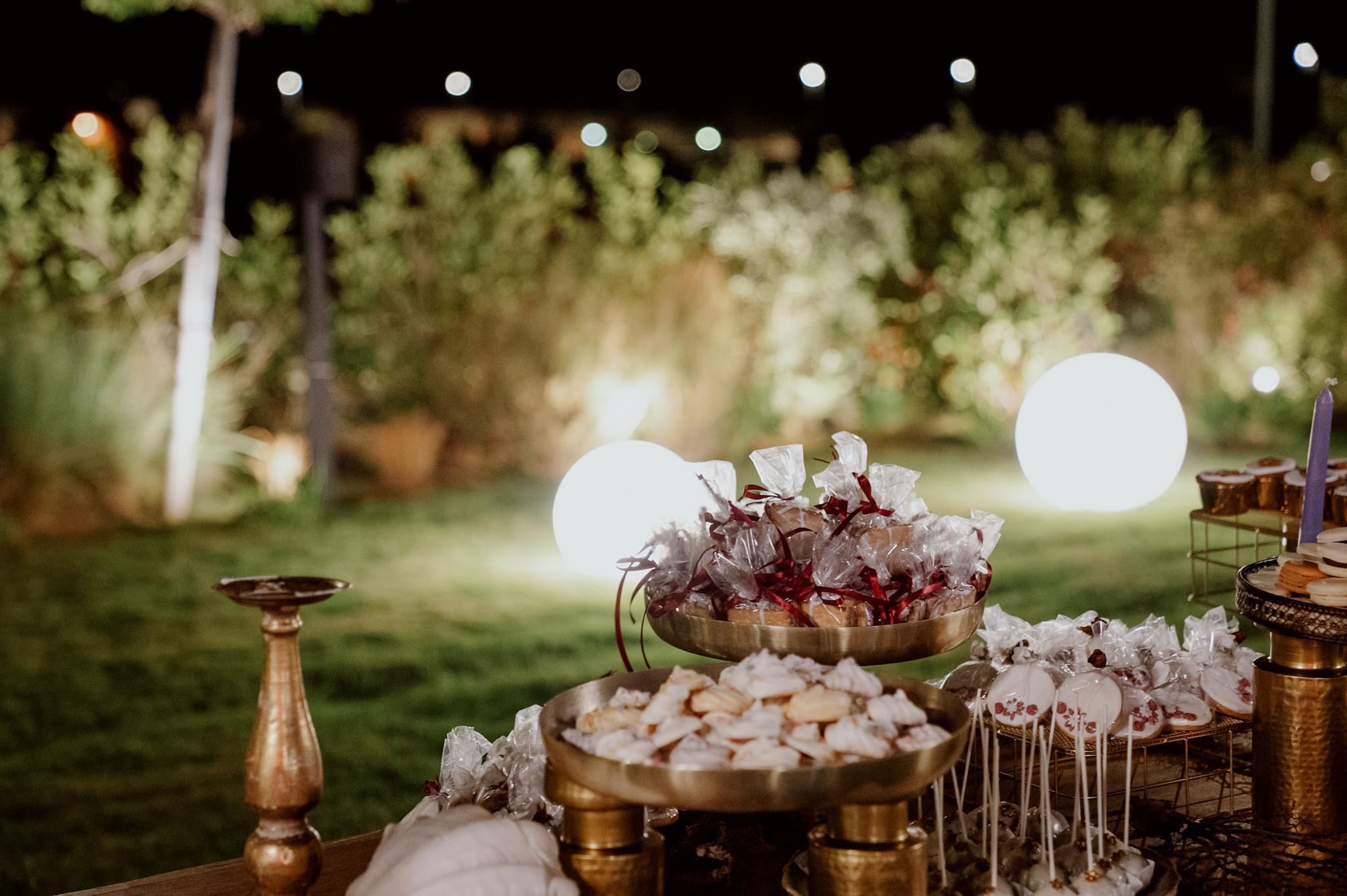 Με φόντο τον πράσινο κήπο και της επιδαπέδιες φωτιζόμενες μπάλες φαίνεται το candy bar το οποίο είναι σε χρωματισμούς του χρυσού και επάνω στα χρυσά σκεύη βρίσκονται cookies μαζί με μαρέγκες και cake pops