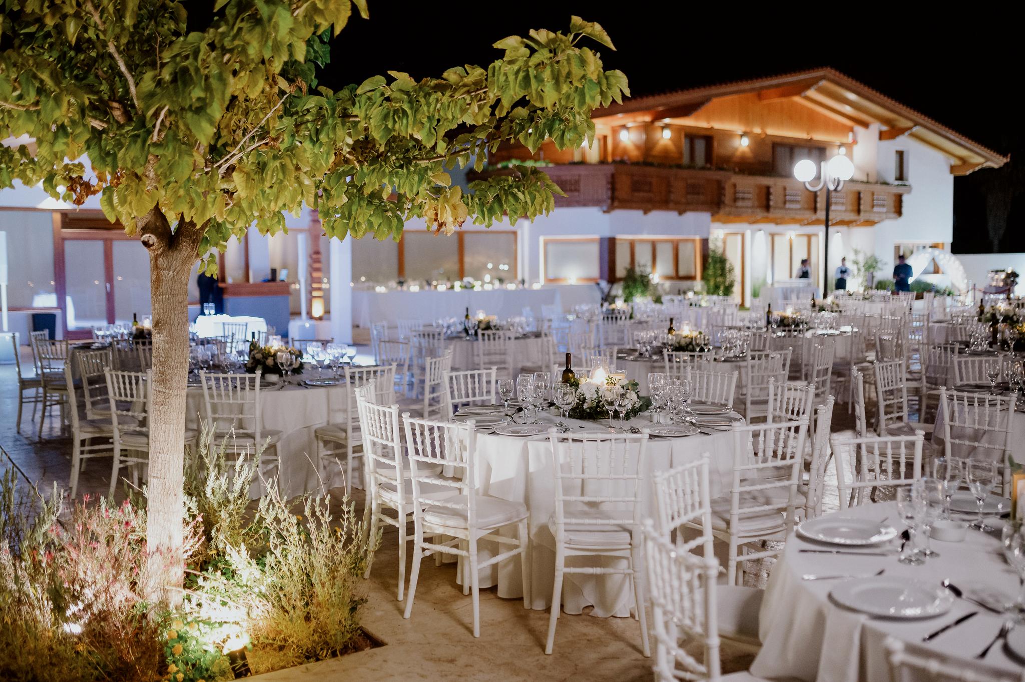 Με φόντο το ελβετικό σαλέ και τις ξύλινες λεπτομέρειες έχουν στρωθεί στον προαύλιο χώρο του κτήματος ροτόντες τραπέζια με περιμετρικά καρέκλες. Στην μέση του κάθε τραπεζιού βρίσκετε μια σύνθεση από ορχιδέες και λευκά τριαντάφυλλα. Επίσης βρίσκετε στην μέση του κάθε τραπεζιού βρίσκετε ένα μπουκάλι κρασί.