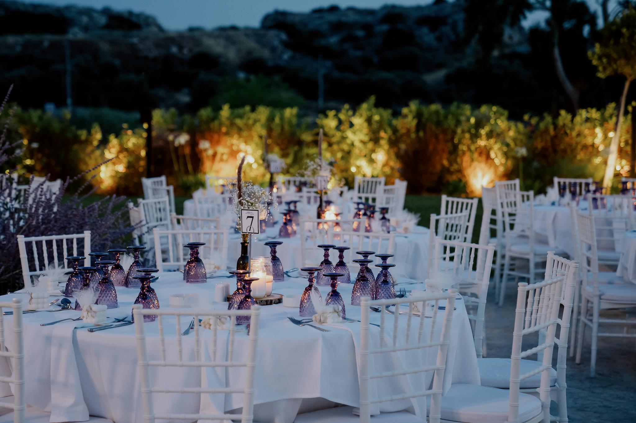 Φωτογραφία με φόντο τον πράσινο κήπο του κτήματος. Μπροστά φαίνονται οι ροτόντες τραπέζια στρωμένες και καρέκλες περιμετρικά από τα τραπέζια.