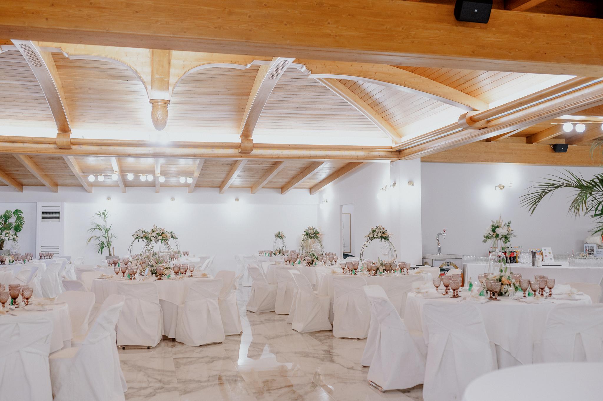 Απομακρυσμένη φωτογραφία στον εσωτερικό χώρο με ροτόντες και τραπέζια περιμετρικά από τις ροτόντες. Επάνω τα τραπέζια είναι στρωμένα με ποτήρια και μαχαιροπίρουνα. Στην μέση των τραπεζιών υπάρχει στολισμός όποιός αποτελείτε από γαρδένιας μαζί με λευκά και σομόν τριαντάφυλλα όπως και πρασινάδες. Στην μέση από το στρογγυλό που αποτελούνται κρέμεται ένα κεράκι . Στο βάθος φαίνονται οι τζαμαρίες του κτήματος που αφήνουν να δεις των έξω χώρο. Φαίνεται η ξύλινη στέγη στην οροφή που υπάρχει