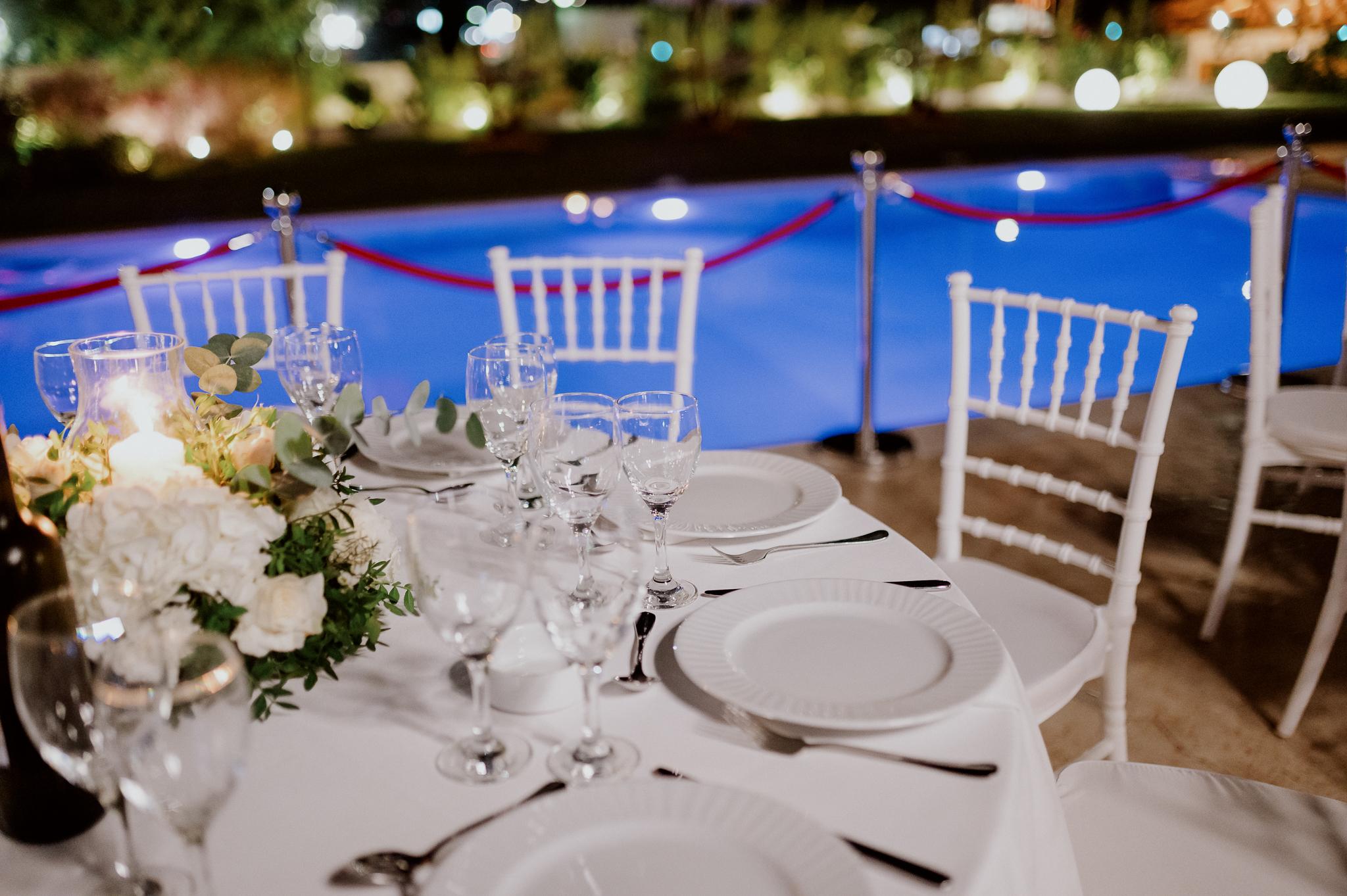 Κοντινή φωτογραφία που στο φόντο φαίνεται η πισίνα. Φαίνεται η μισή ροτόντα με στρωμένο το τραπέζι και τα πιάτα . Στολισμός τραπέζιου βρίσκετε στην μέση και περιέχει τριαντάφυλλα λευκά με ένα κερί στην μέση