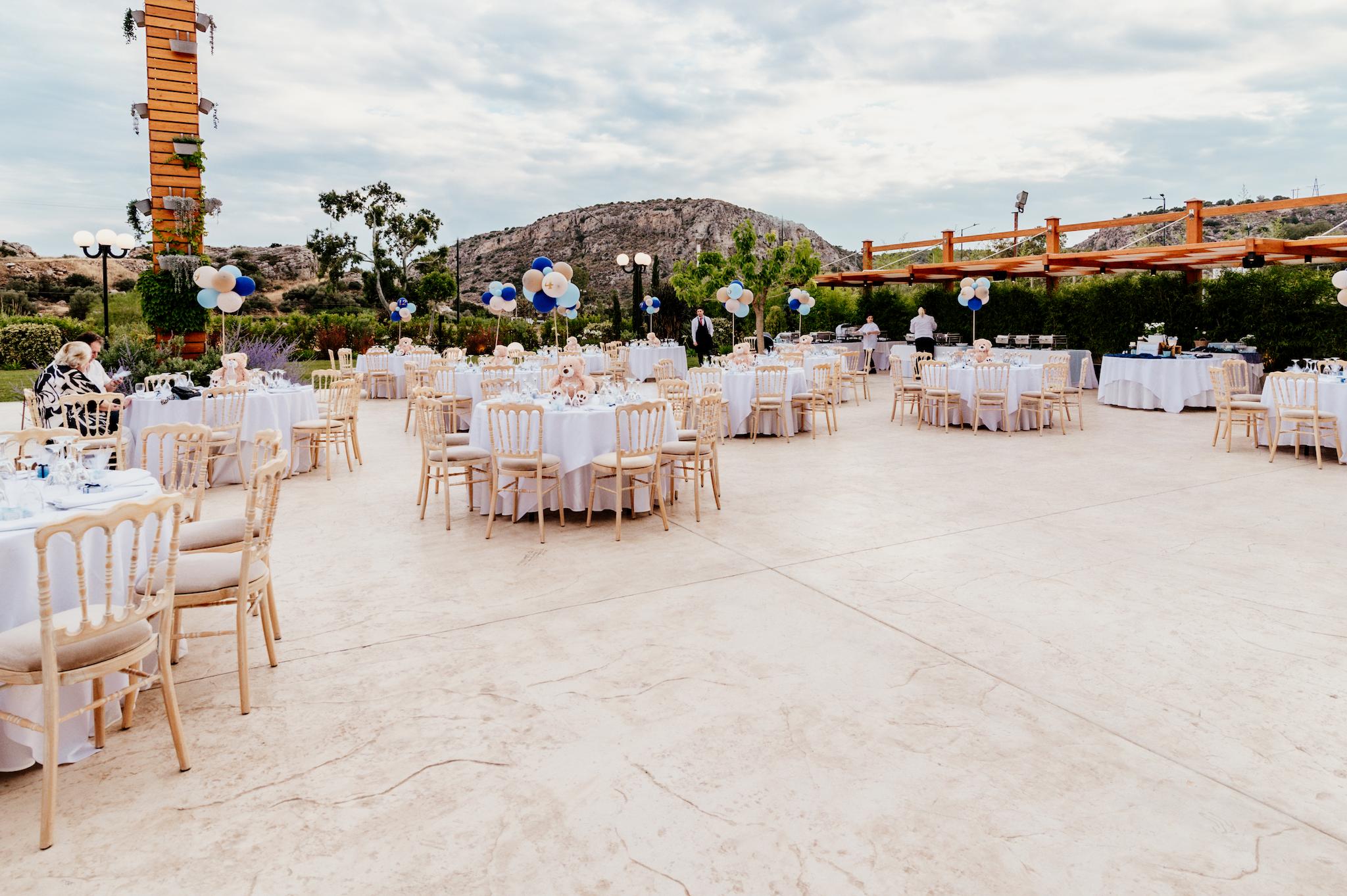 Απομακρυσμένη φωτογραφία ο εξωτερικό χώρος του κτήματος στον οποίο τα τραπέζια είναι στημένα από το catering. Υπάρχουν ροτόντες με περιμετρικά ξύλινες καρέκλες. Στο κέντρο του κάθε τραπεζιού υπάρχει στολισμός με ένα αρκουδάκι το οποίο κρατάει σύνθεση από μπαλόνια. Στο βάθος βρίσκετε το πράσινο τοπίο αλλά και η πέργκολα που είναι από κάτω το catering